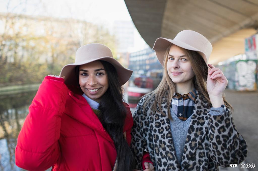 To jenter i forskjellige antrekk. Den ene har rød ytterjakke, den andre jakke med leopardmønster. Begge har bredbremmet hatt. Foto. 