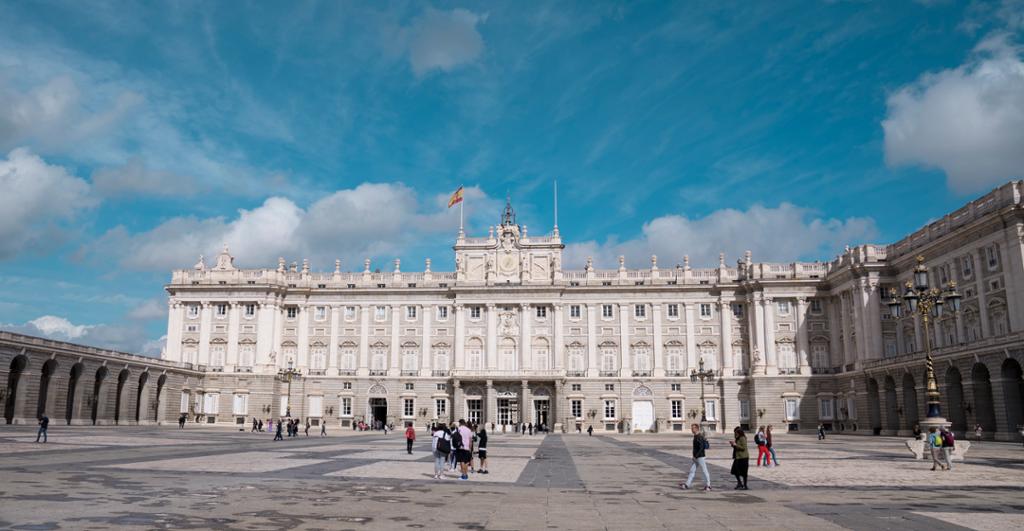 Et stort palass rundt en åpen plass hvor mennesker rusler rundt. Det spanske flagget vaier over bygget. Foto.