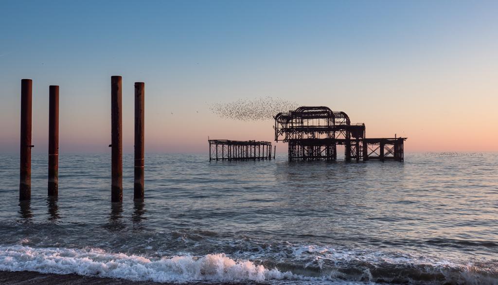 Photo of the ruins of an old pier in England. The sun is setting so the sky is blue and pink. We see the ocean. 