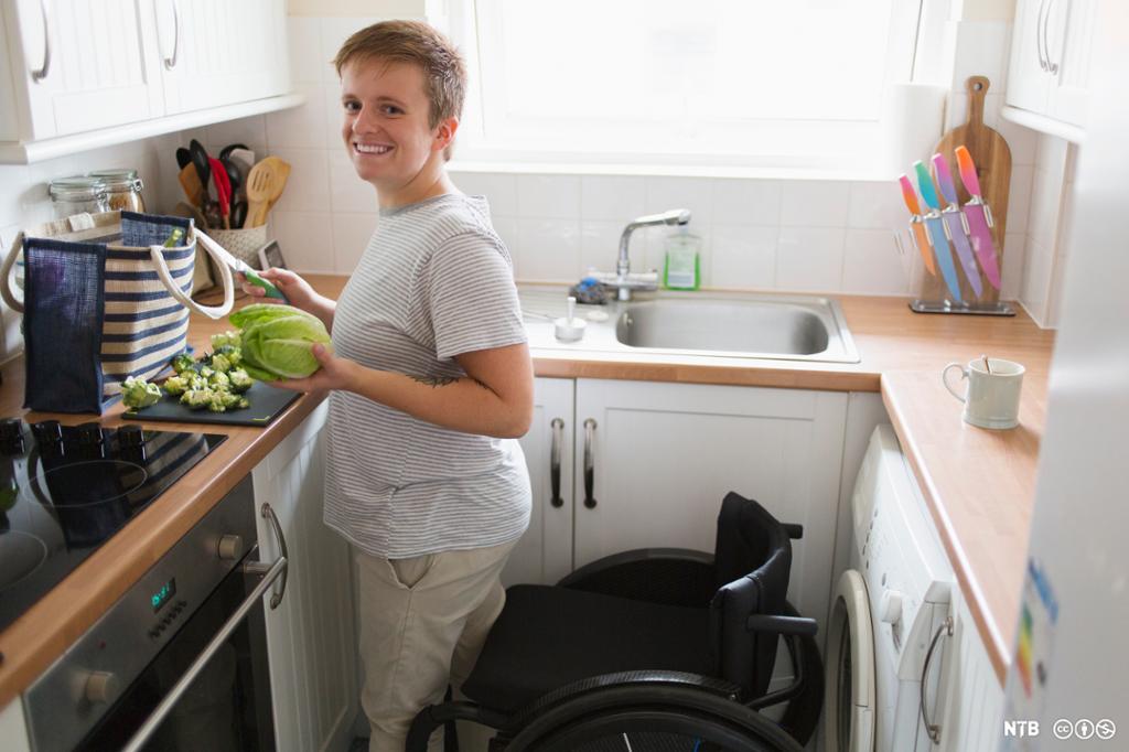 Ung kvinne lager mat ved kjøkkenbenken mens hun står foran rullestolen sin. Foto.