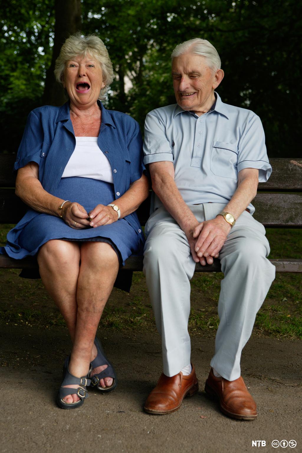 Et eldre par sitter på en benk i en park. Damen ler og mannen smiler. Foto.