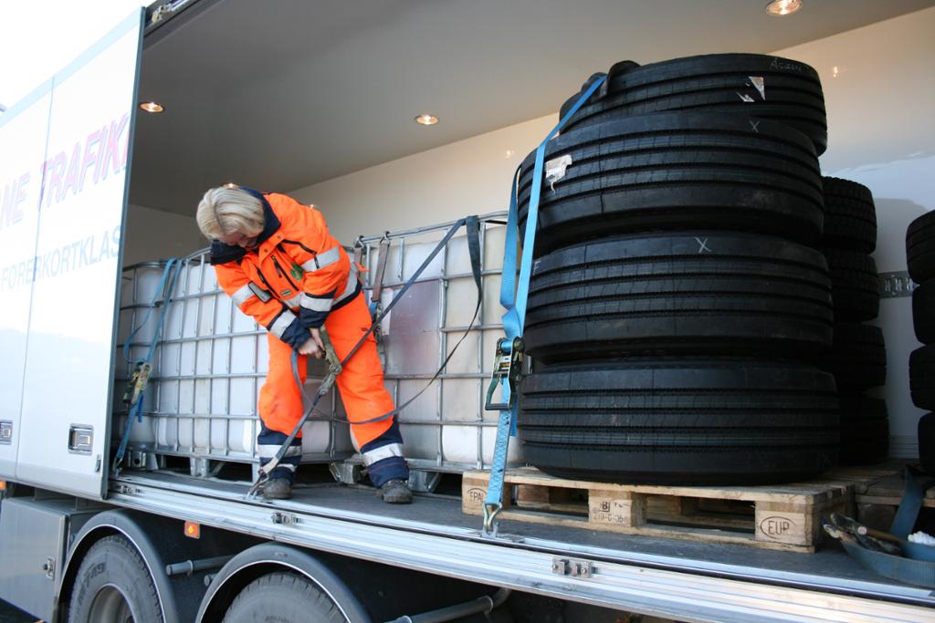 En mann i oransje arbeidsklær sikrer lasta på en lastebil med fiberstropper. Foto.