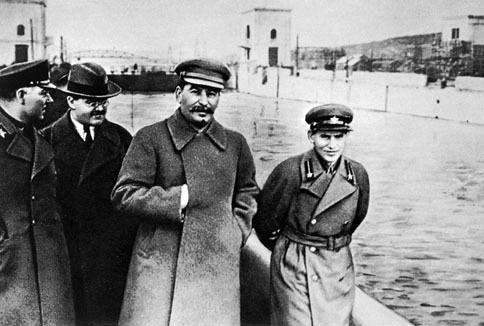 Josef Stalin med tre andre sentrale kommunistledere går langs elva. Mennene har ulike hatter og frakker. Stalin har den ene hånda innenfor frakken. Foto.