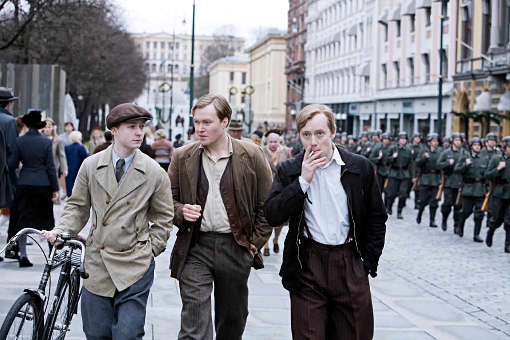 Foto av tre unge norske menn med sykkel, sigaretter og klær fra 1940-tallet. De spaserer nedover Karl Johans gate i Oslo mens tyske tropper marsjerer inn i byen.