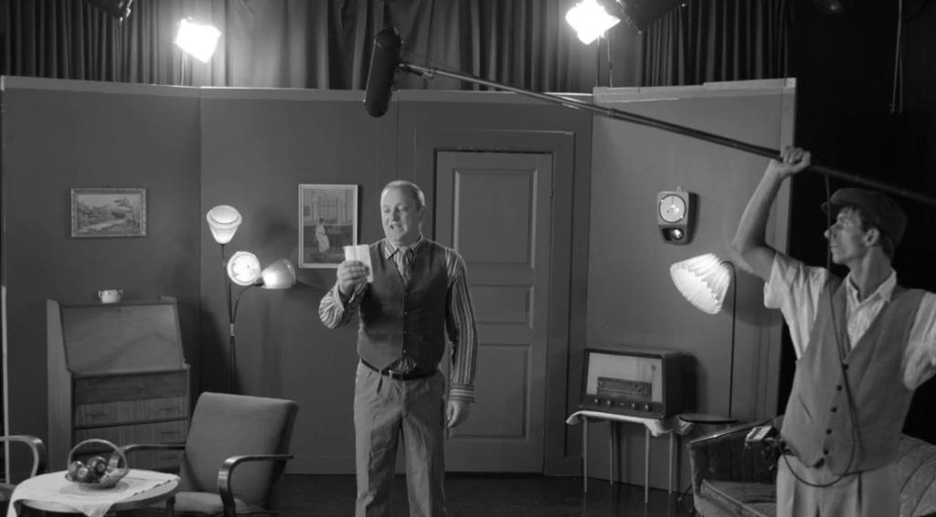 Sort/hvitt-bilde av en mann som står i et rom og snakker. Ved siden av står en mann med lang mikrofon og spiller inn lyden. Foto.