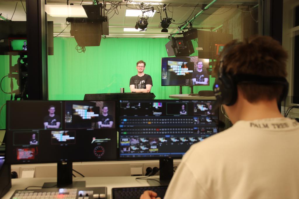 I et TV-studio med grønnskjerm, lyskastere og kamera står en programleder. I forgrunnen ser vi ryggen til en ung mann som følger med på skjermene i kontrollrommet. Foto.