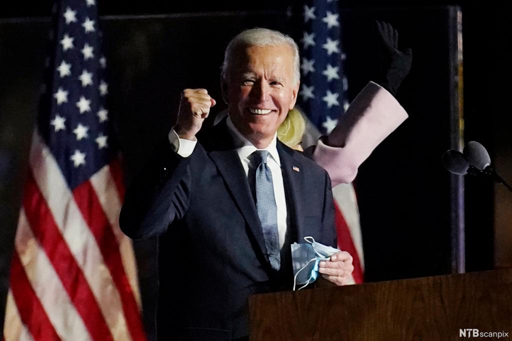 Photo: We see Joe Biden smiling while clenching his right fist. He is standing in front of American flags. He is a grey-haired man in a dark suit. 