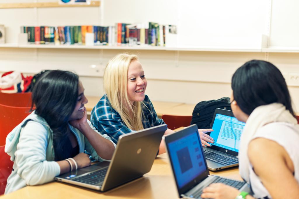 Tre elever sitter rundt et bord med laptoper og prater. Foto.