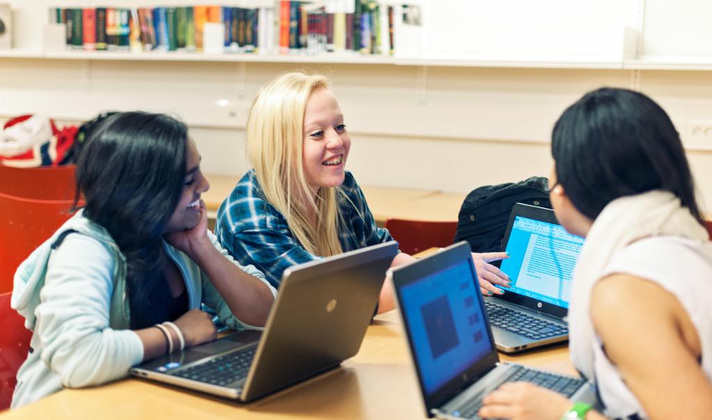 Photo: Three girls are talking and working together. Each girl has a computer. 