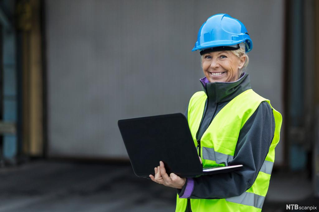 Smilende kvinne iført hjelm og synlighetsvest står utendørs med en åpen laptop i hendene. Foto.