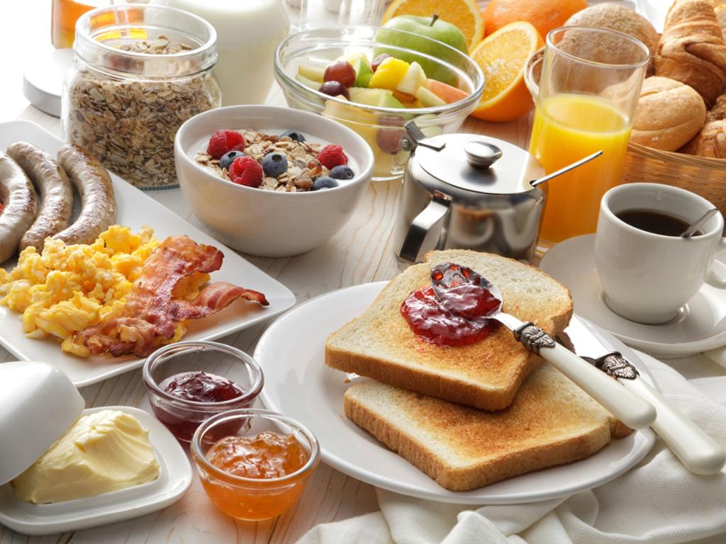 Et stort frokostbord med eggerøre, bacon og pølser, syltetøy, smør, ulike brødvarer, oppkuttet frukt, müsli, kaffe, melk og appelsinjus. Foto.