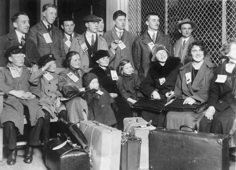Black and white photo: We see a group of immigrants seated. They are men, women and children. They have white cards tacked to their collars. We also see suitcases in the picture. 