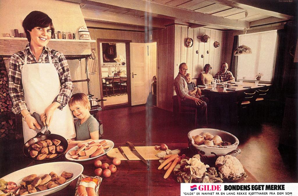 Reklame for Gilde fra 1977. En kvinne med forkle tilbereder og serverer en rekke kjøttvarer fra Gilde. Et lite barn står inntil henne og smiler. Ved et dekket bord i bakgrunnen sitter to menn, en kvinne og et barn. Et tredje barn står i en døråpning. Foto. 