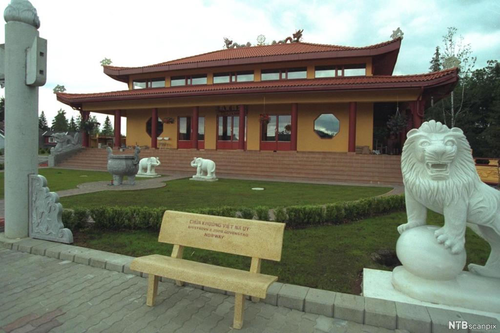 Gul bygning med pagode-tak. Skulptur av løve og en benk i forgrunnen. Foto.