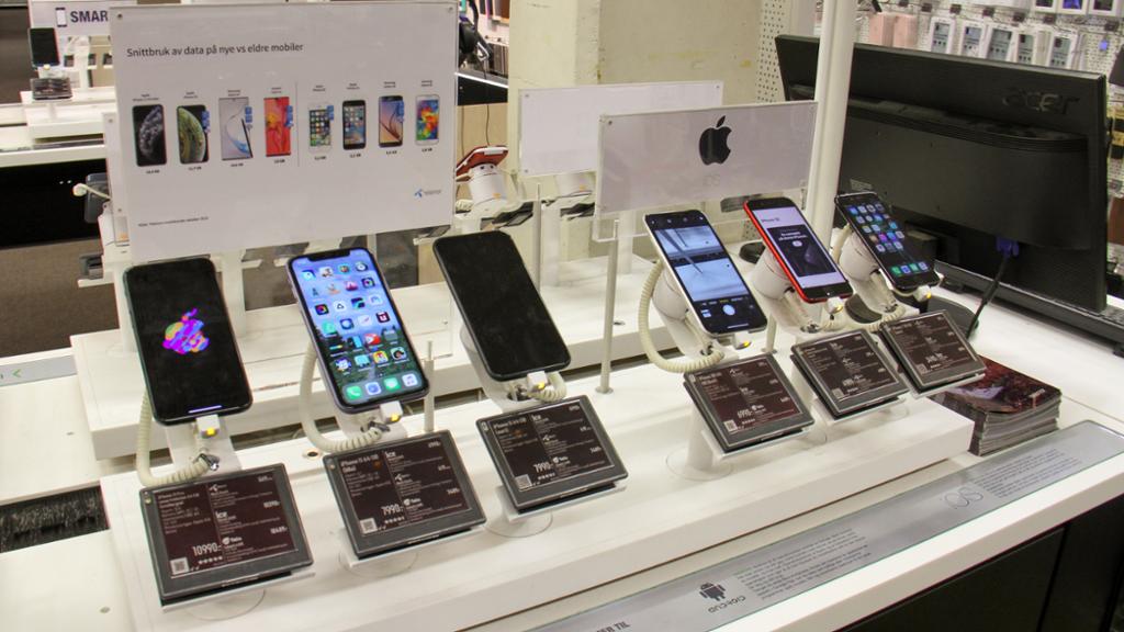 Utstilling av ulike modeller av iPhone. De står utstilt etter hverandre på ei hylle. Over dem henger en plakat med informasjon om telefonene. Foto.