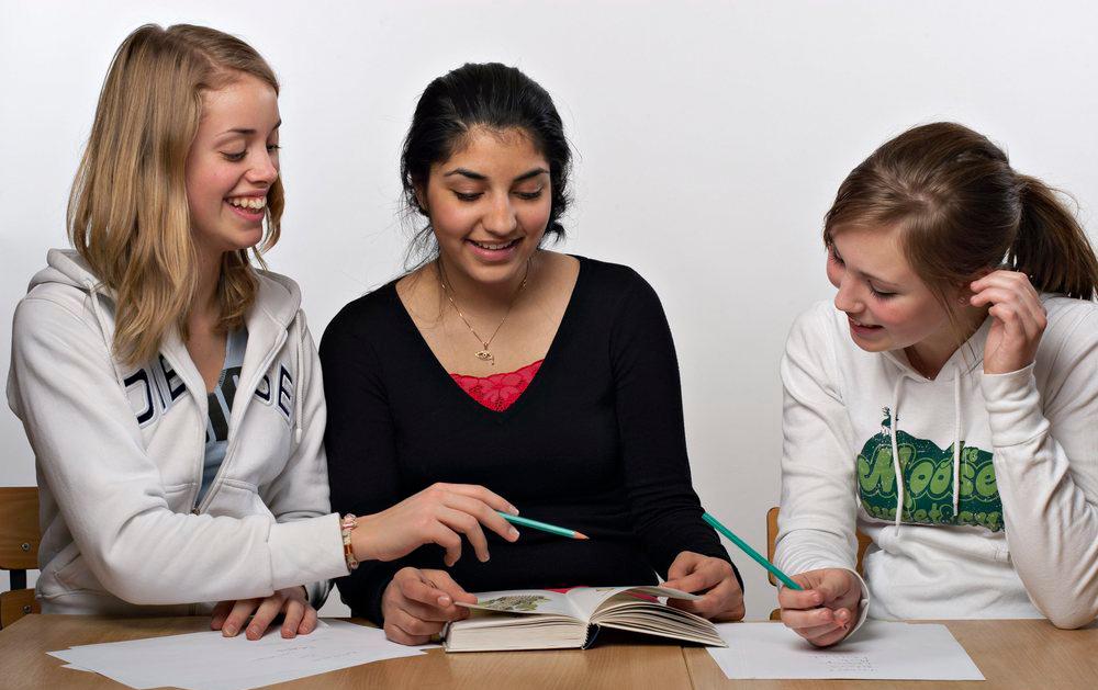 Tre unge jenter sitter bak en pult. De har ei åpen bok og flere ark foran seg, og den ene peker med en blyant på noe i boka. Alle smiler. Foto. 