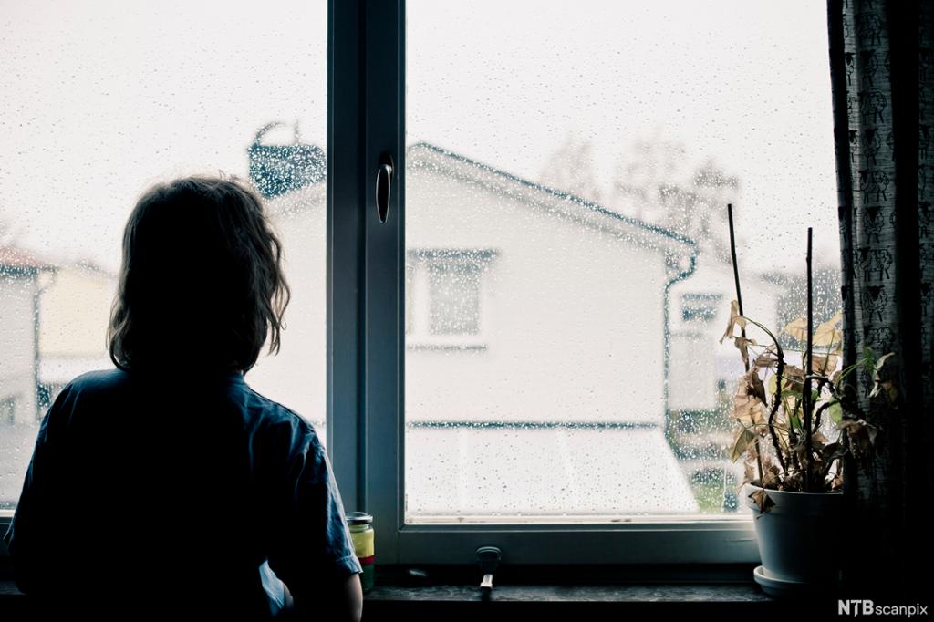 En ung jente ser ut av et vindu med regn på. Vi ser hus i bakgrunnen. Foto. 