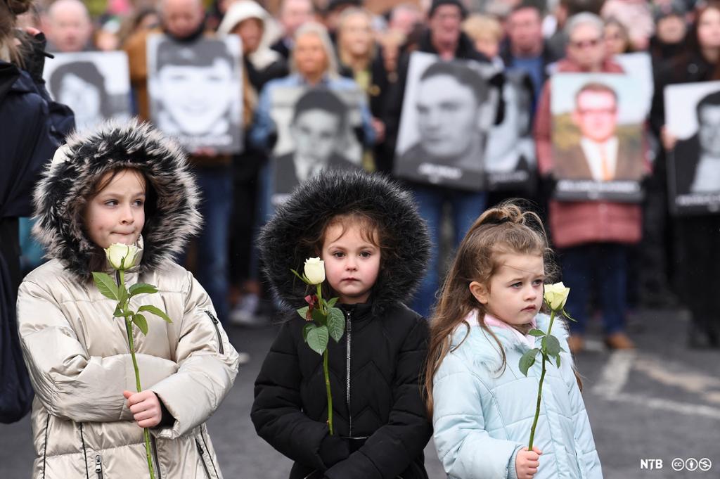 Photo: We see three children holding white roses. Behind them, we see people holding up photos of the victims of the Bloody Sunday massacre. 