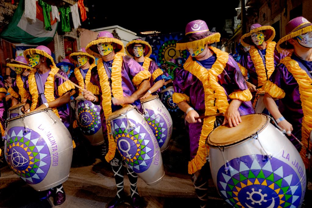 Flere personer i fargerike kostymer spiller på store trommer mens de går i gata. Foto.