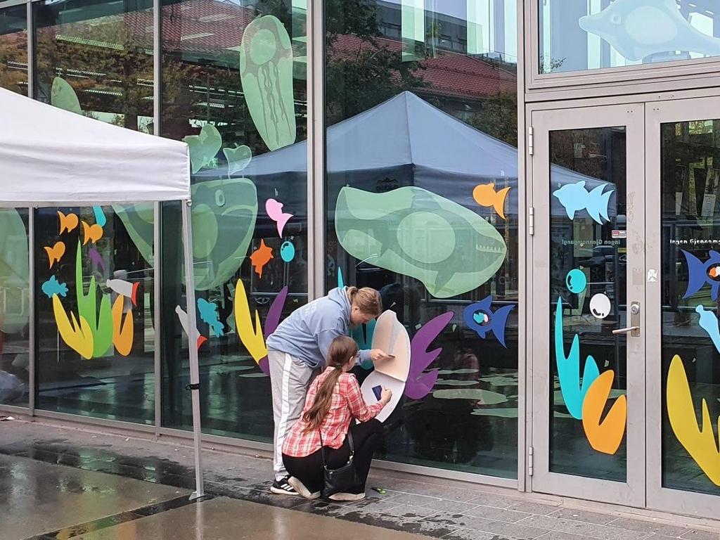 Skuleelevar festar fargerike dekorasjonar på glasveggane til Asker kulturhus. Foto.