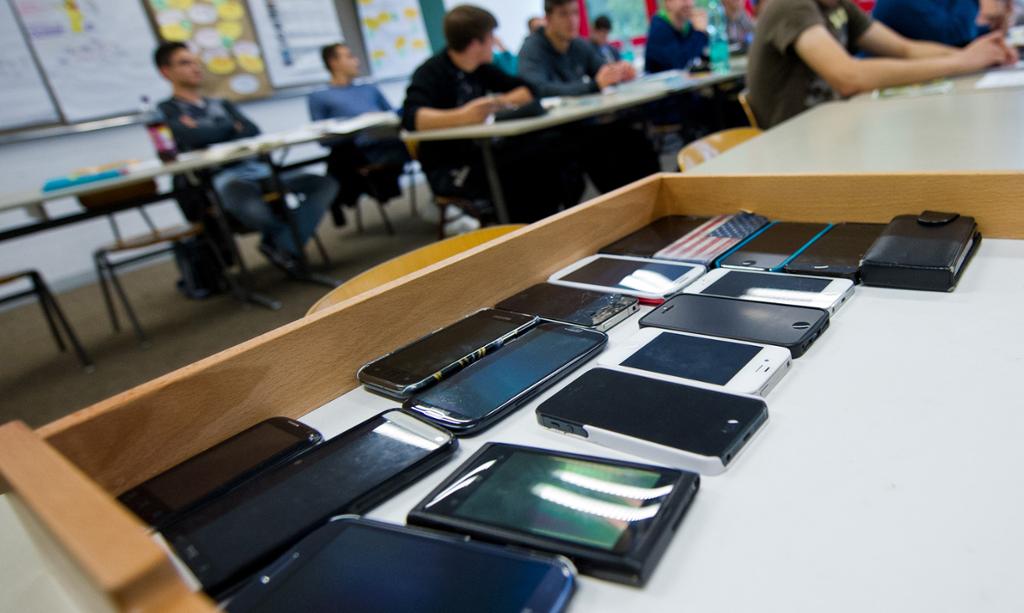 17 mobiltelefoner som ligger samla på en pult i et klasserom; elever i bakgrunnen. Foto.
