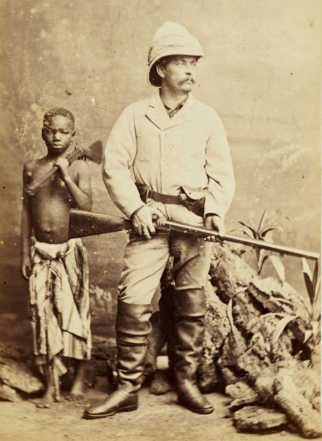 Henry Morton Stanley, britisk journalist og oppdager i Afrika, sammen med sin personlige tjener og adopterte sønn Kalulu. Kalulu står i bakgrunnen. Stanley poserer med gevær. Foto.