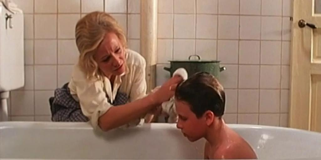 En gutt sitter i badekaret. En kvinne sitter bøyd på gulvet ved badekaret og vasker håret hans. Foto. 