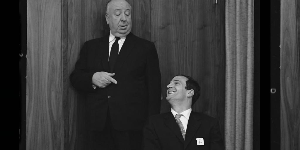 En eldre mann, Alfred Hitchcock, står og peker ned på en yngre leende mann som sitter ved siden av ham, François Truffaut. Begge har på seg mørk dress. Svart-hvitt foto.