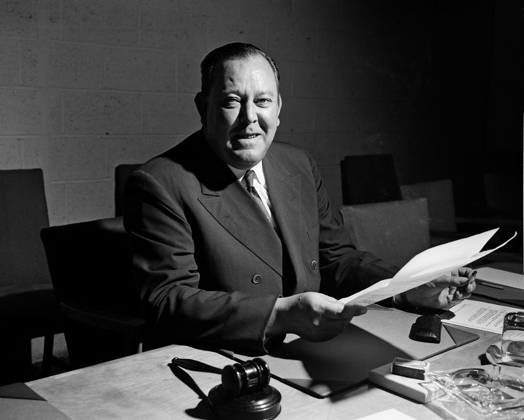 Mann med dress og slips sitter ved et skrivebord med ark i hendene og en klubbe ved siden av seg. Svart-hvitt foto.