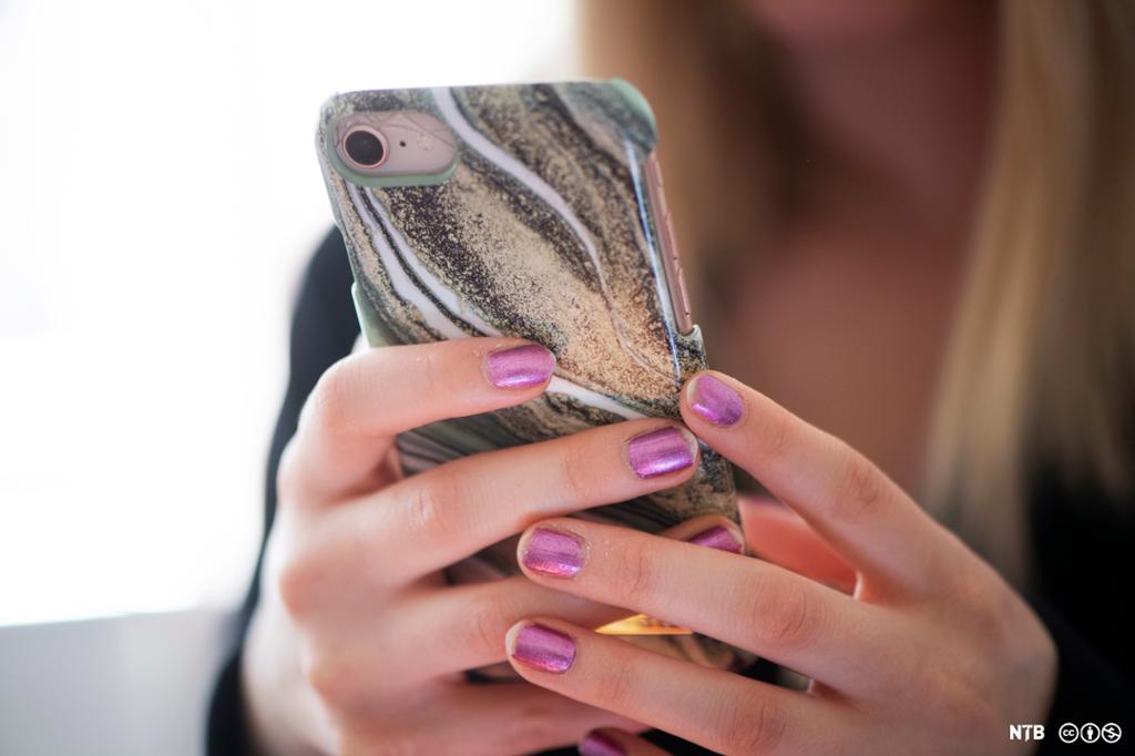 Tenåringsjente holder en mobiltelefon i hendene. Neglene er malt med lilla neglelakk. Foto.