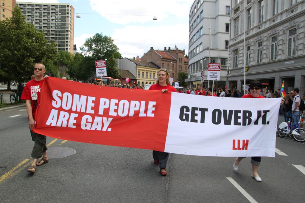 Tre personar som held ein plakat i samband med pride-paraden med teksten: "Some people are gay. Get over it." Foto.