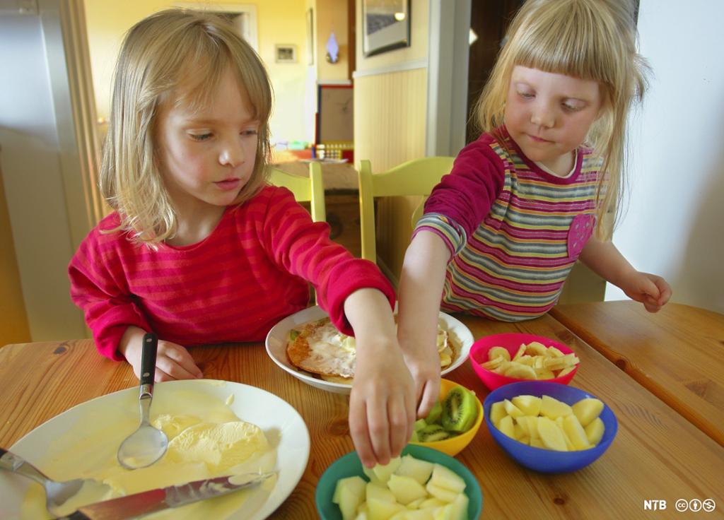 To små jenter sitter ved et bord og forsyner seg med hendene av oppskåret frukt fra skåler foran seg. Foto.