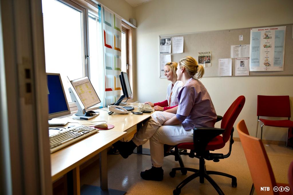 To helsefagarbeidarar sit på rapportrommet og jobbar saman på ein PC. Foto. 
