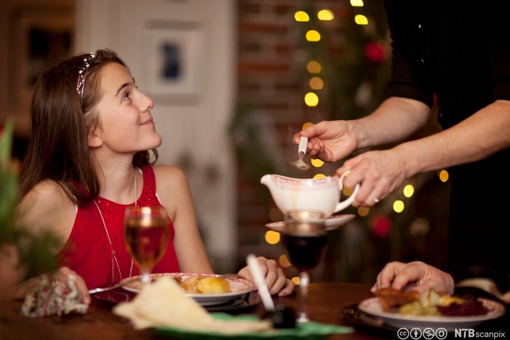Ei ung jente sitter pyntet ved bordet og ser smilende opp på en person som serverer saus. Foto.