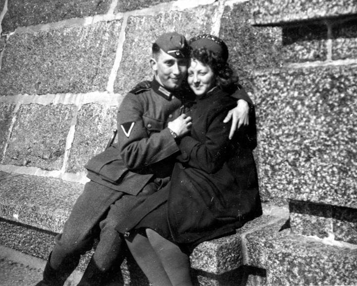 En mann i soldatuniform og ei kvinne med ikledd tykk kåpe og hatt sitter på en steinbenk. Svart-hvitt foto.