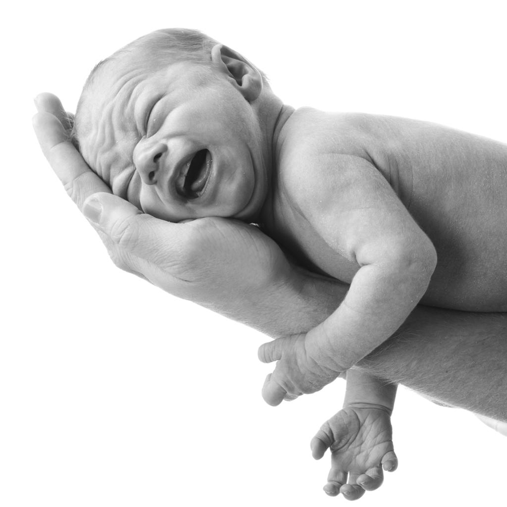 Nyfødt baby som ligger på armen til en voksenperson. Hodet hviler i handflata på den voksne. Babyen har rynket panna og knepet igjen øynene. Foto.