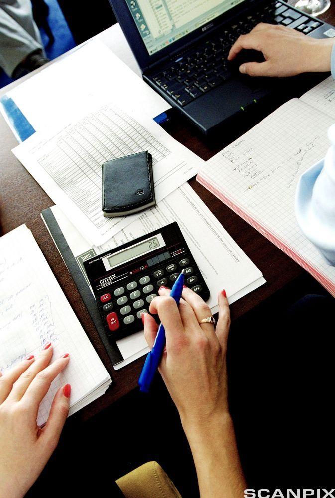 En hånd hviler på et papir, mens en hånd trykker på en kalkulator. Foto.