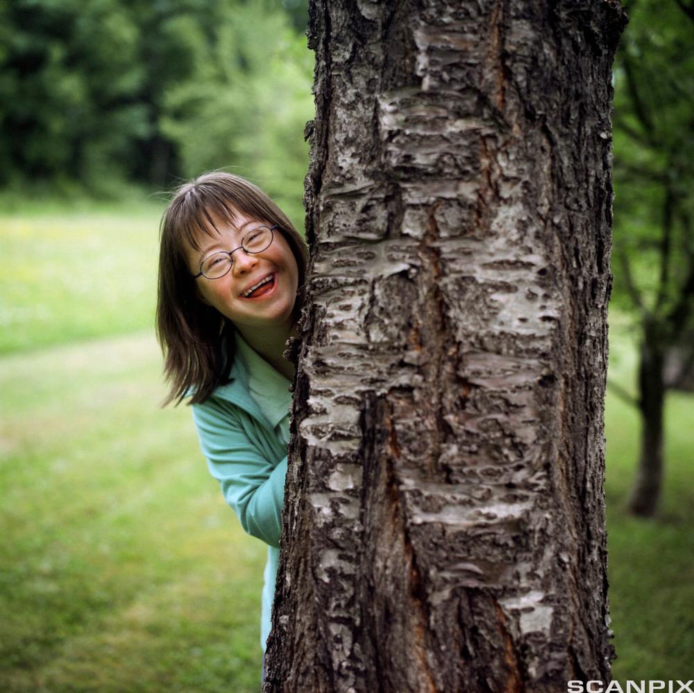Jente med Downs syndrom står bak et tre. Foto. 