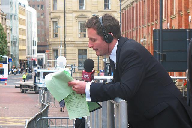 En dresskledd mann med hodesett og mikrofon lener seg over et metallgjerde og leser opp fra en notatblokk. Foto.