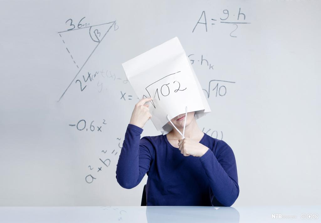 En elev sitter foran en hvit tavle med en papirpose over hodet. På tavla og papirposen er det skrevet ulike matematiske formler og figurer. Foto.