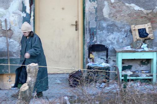 Dame i slitte klær foran et forfallent hus med søppel og slitte møbler utenfor. Foto.