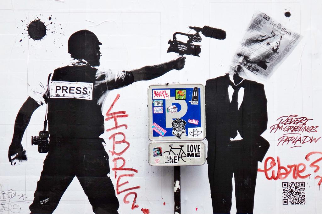 Grafitti av en pressemann som bruker et videokamera til å skyte ei avis på en dresskledd mann. Foto.