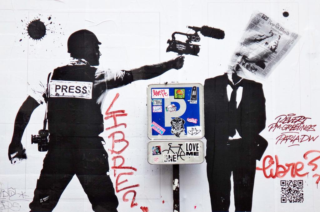 Grafitti av en pressemann skyter en avis på en dresskledd mann med videokamera. Foto.