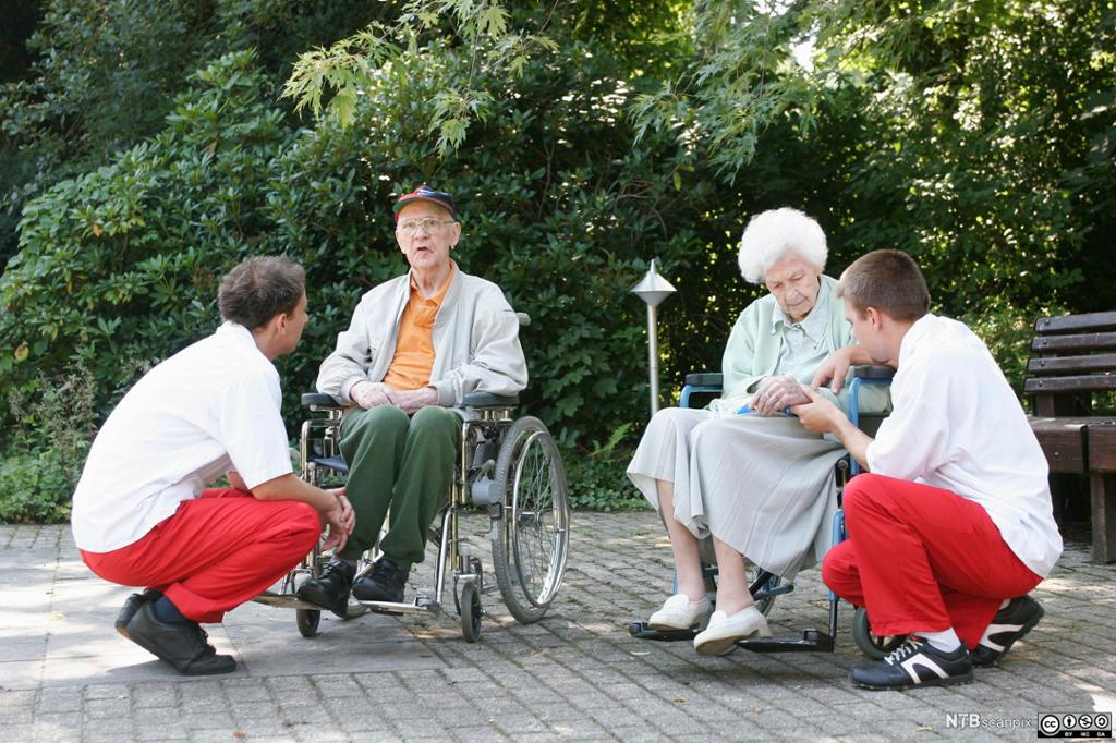 To eldre ute i rullestol kommuniserer med to helsefagarbeidere. Foto.