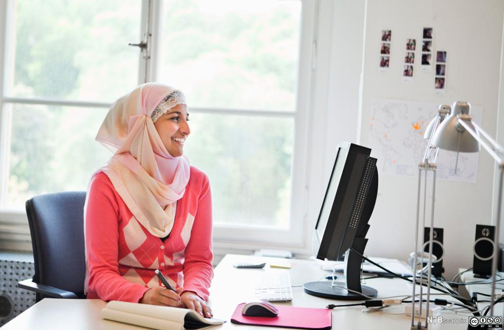 Ung jente i hijab sit framfor ei datamaskin og tek notat. Foto.