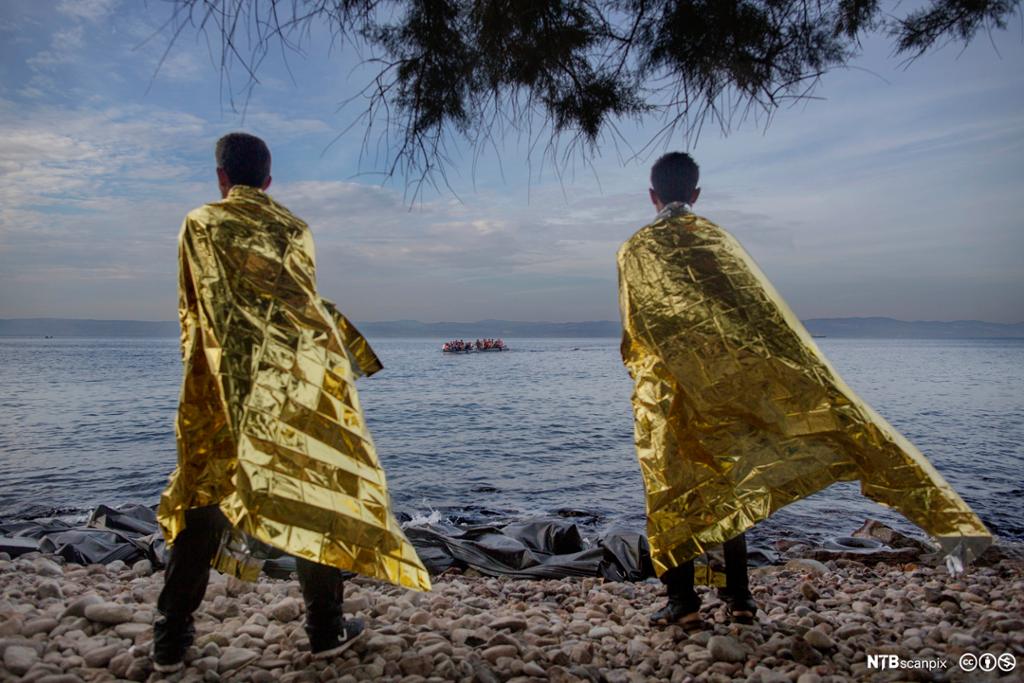 To menn innpakket i folie for å få varme i kroppen ser en ny båtlast med flyktninger i gummibåt på vei mot land. Foto.