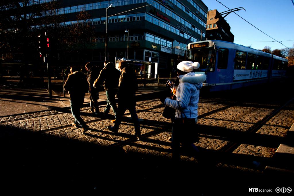 Mennesker i ferd med å krysse en fotgjengerovergang i Stortingsgata ved Nasjonalteateret i Oslo sentrum på rødt lys. En trikk venter på at fotgjengerne krysser gata. Foto.