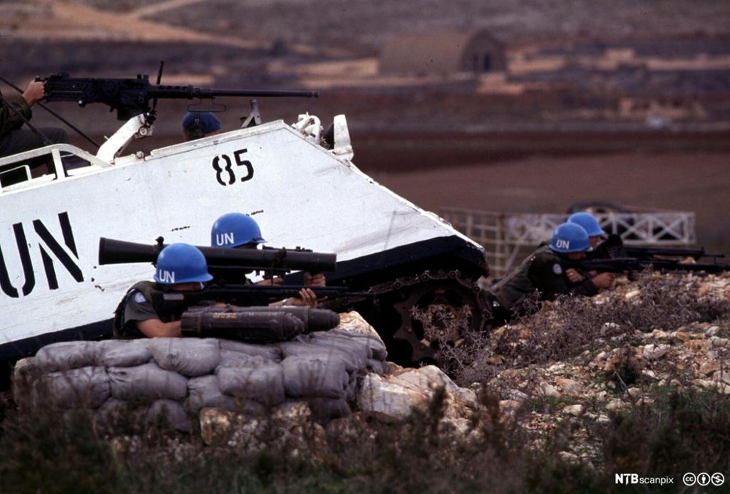 FN-soldater med skytevåpen bak sandsekker ved siden av armert kjøretøy. Foto.