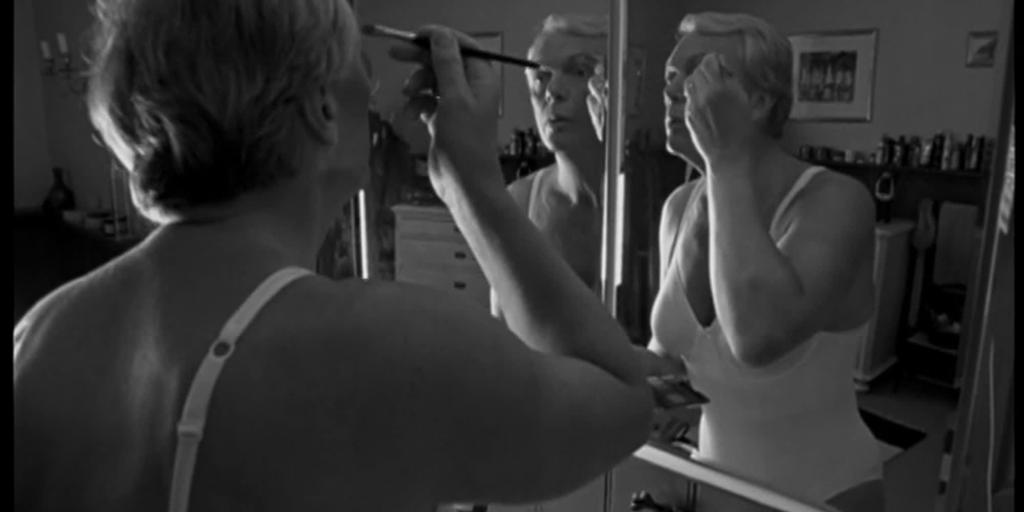 Svart-hvitt-bilde av en transperson med kort, grått hår og kvinneundertøy som står foran speilet og sminker seg. Foto.
