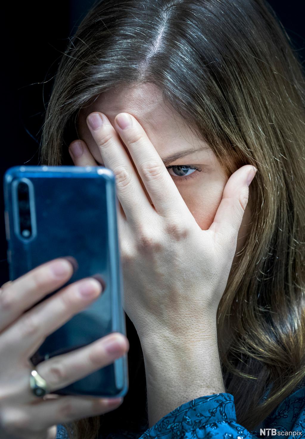 En kvinne holder hånda foran ansiktet mens hun ser på en mobiltelefon. Foto. 